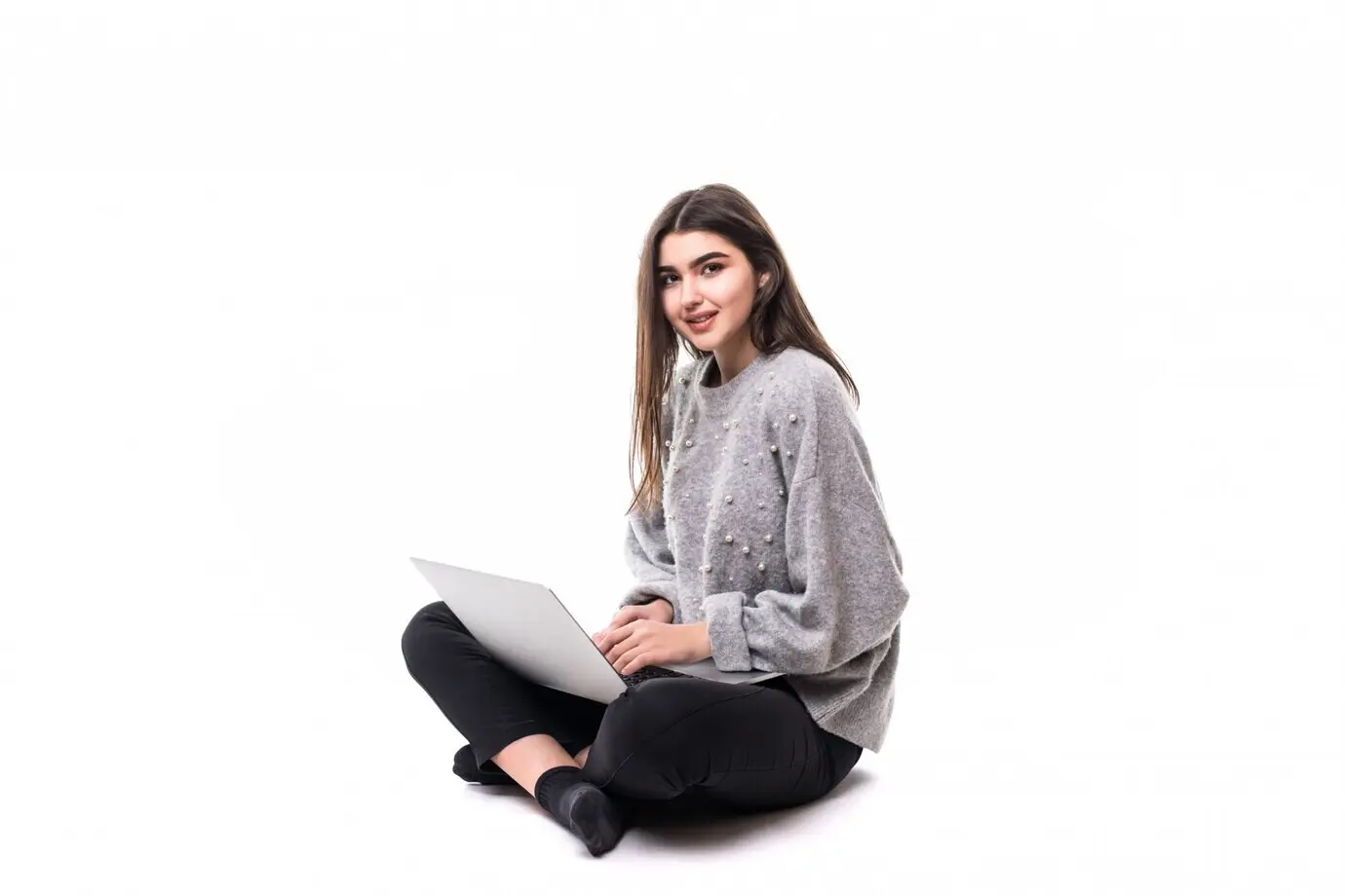 Lächelndes brünettes Mädchen-Model in einem grauen Pullover sitzt auf dem Boden und arbeitet und studiert an ihrem Laptop.